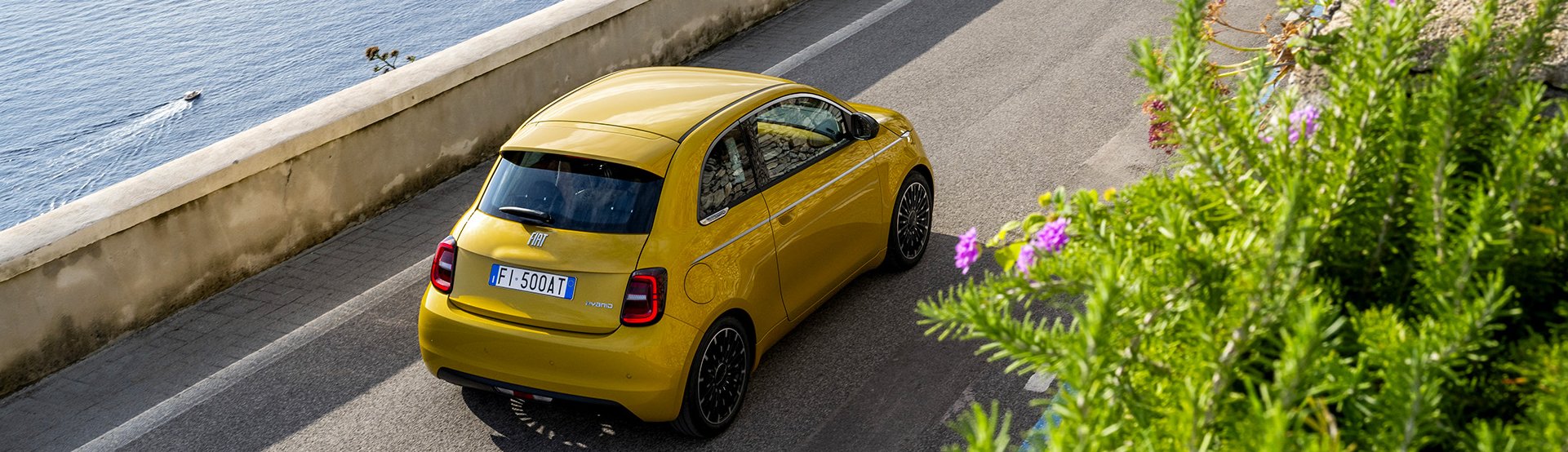 Yellow car driving on coastal road with sea view and vegetation.